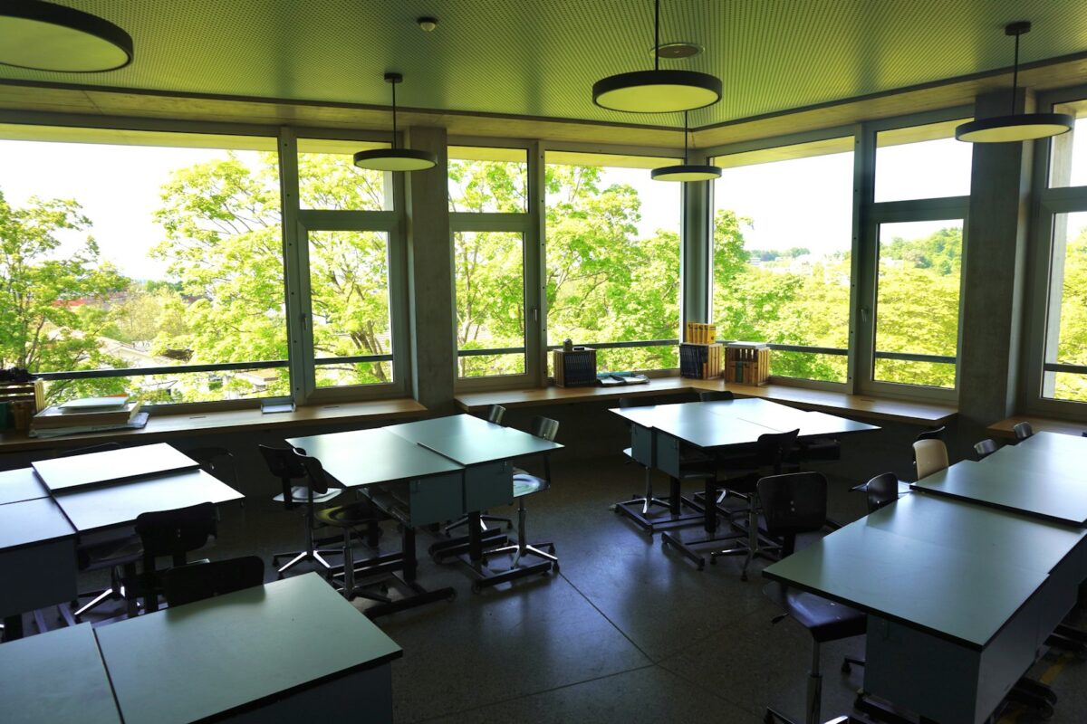 Leeres Schulzimmer mit Pulten und Stühlen vor einer breiten Fensterfront, draussen grüne Baumkronen