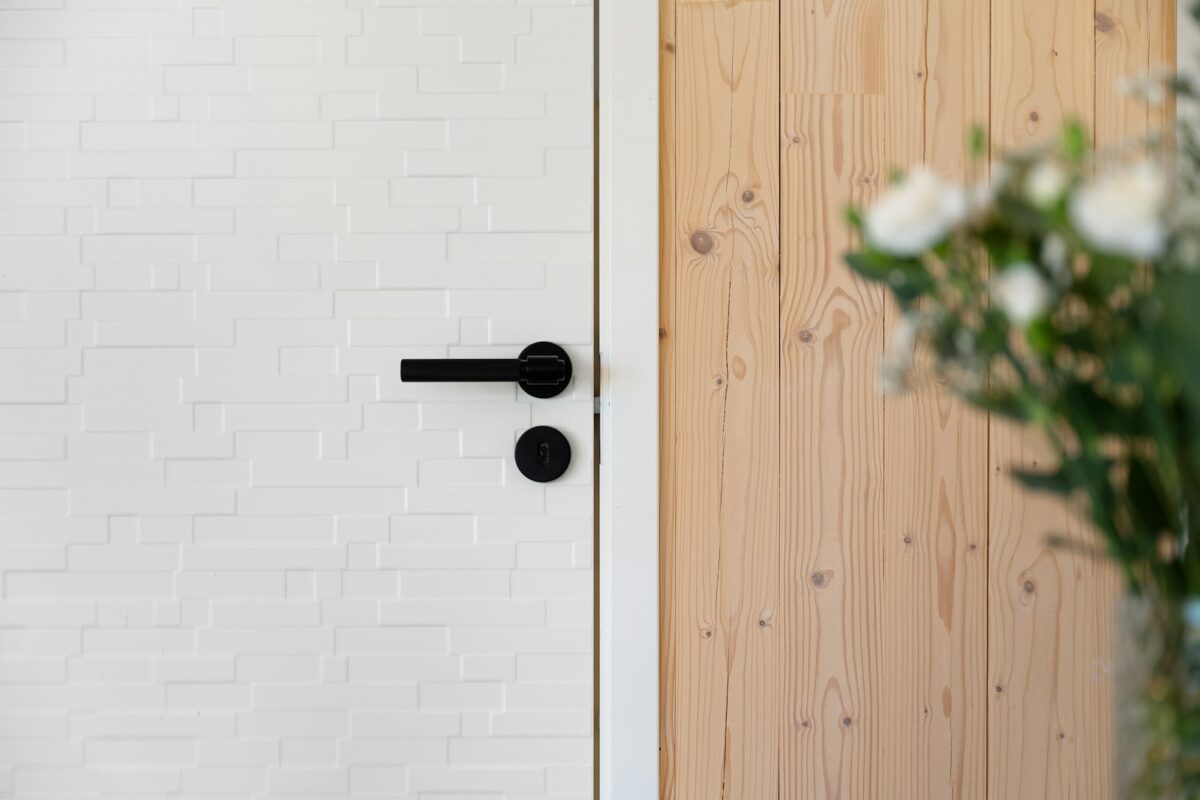 Geschlossene weisse Tür mit schwarzer Klinke neben einer Holzwand, rechts unscharf ein Blumenstrauss